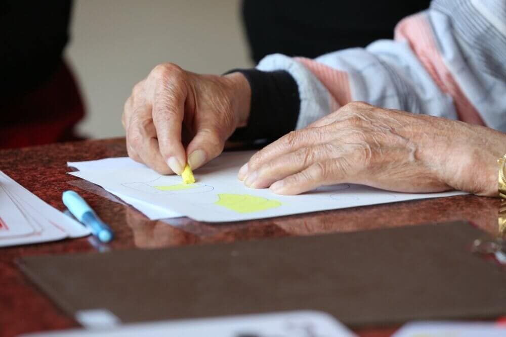 Close-up of elderly hands using a yellow crayon to color on paper on a table. - Home Instead