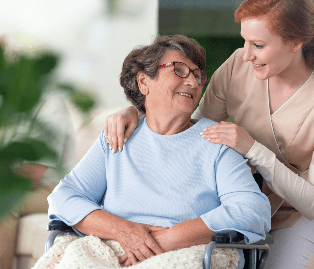 Older woman in a wheelchair smiling at a younger woman who is touching her shoulder and smiling back. - Home Instead
