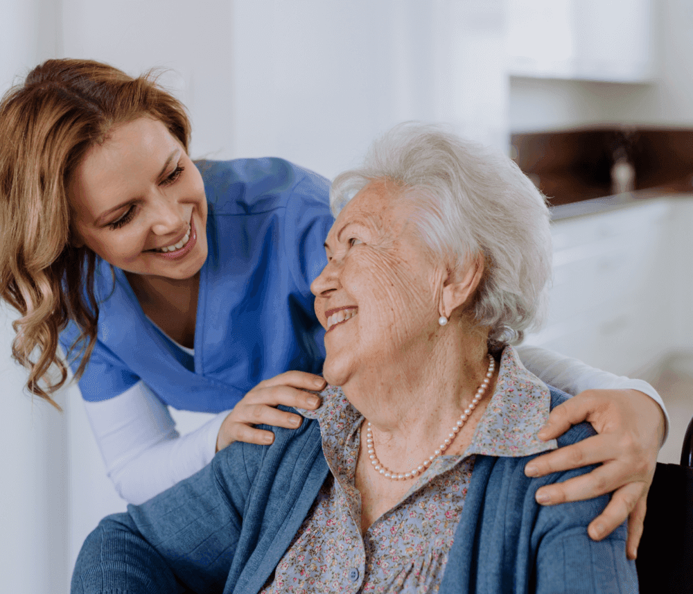 A young nurse smiles and gently leans on the shoulders of an elderly woman in a wheelchair, who is also smiling. - Home Instead