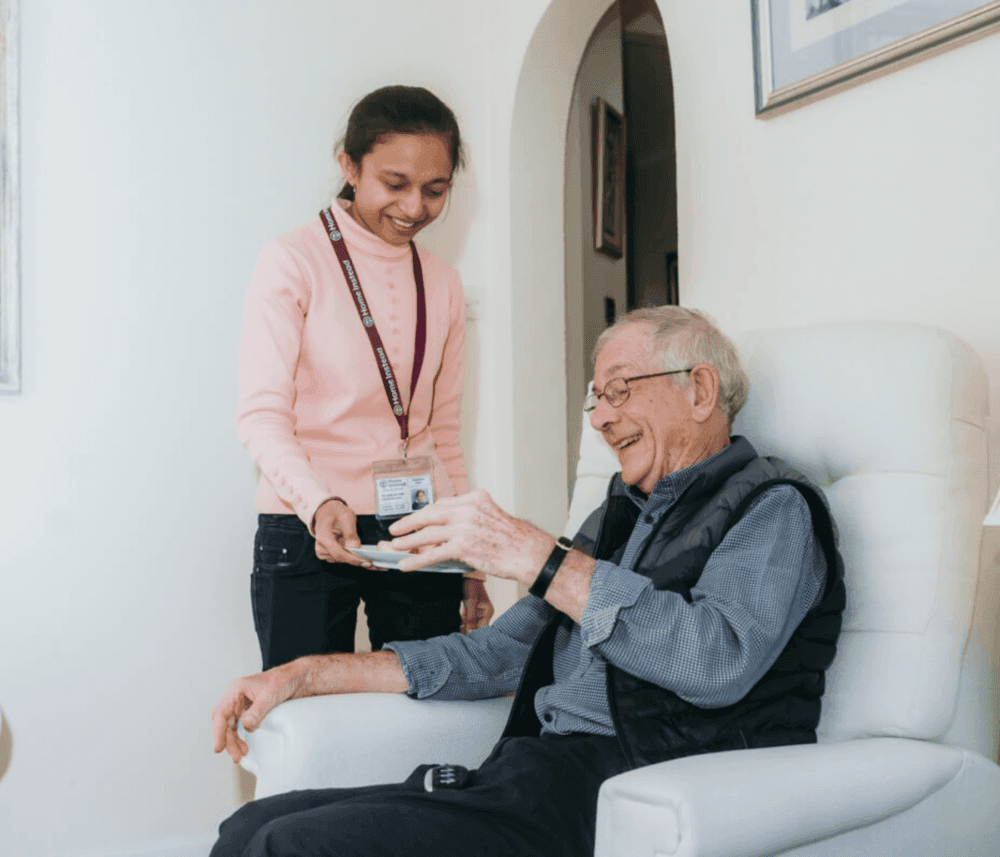 Young woman hands a drink to a smiling elderly man sitting in a white chair inside a cozy, well-lit room. - Home Instead
