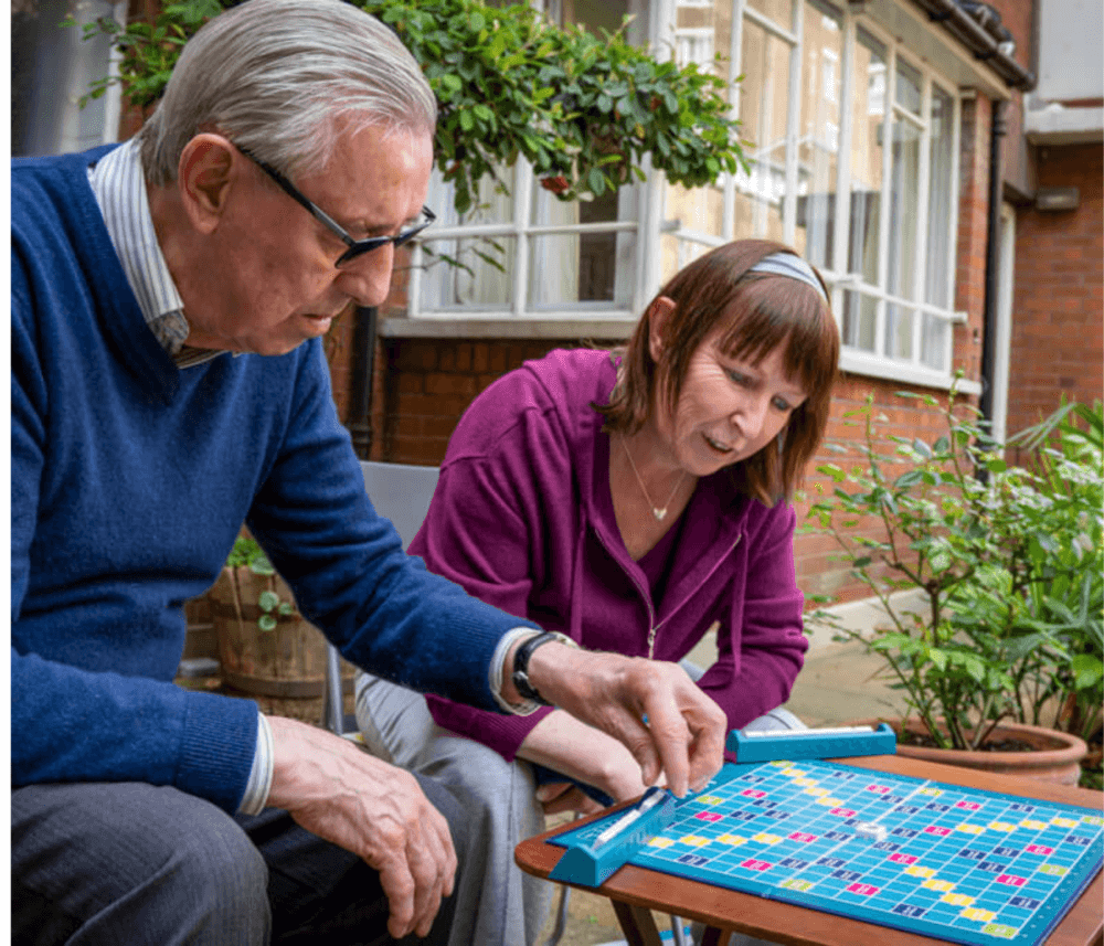 Two people are playing Scrabble outside next to a building, concentrating on arranging their tiles on the board. - Home Instead