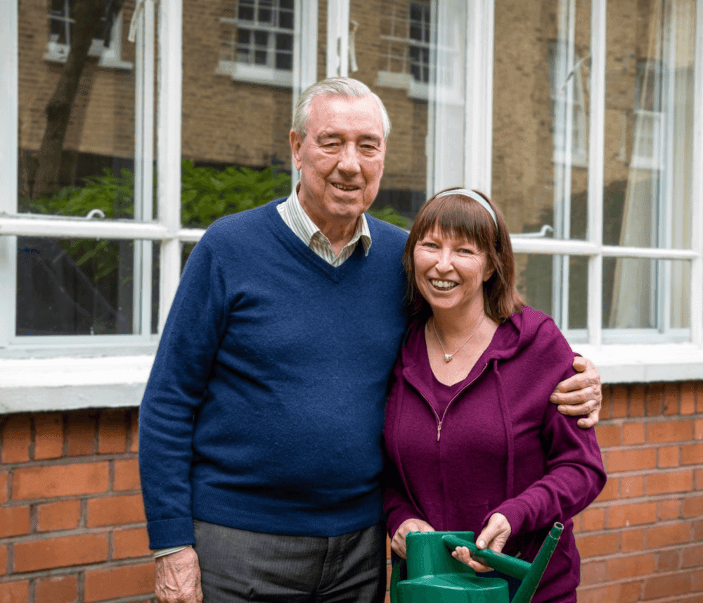 An elderly man and a woman smile while holding a watering can in front of a building with large windows. - Home Instead