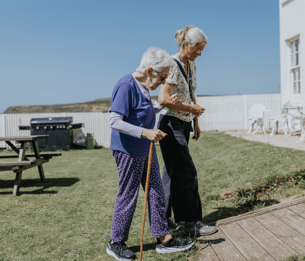 An elderly woman with a cane walks with the assistance of a caregiver outside in a grassy area on a sunny day. - Home Instead