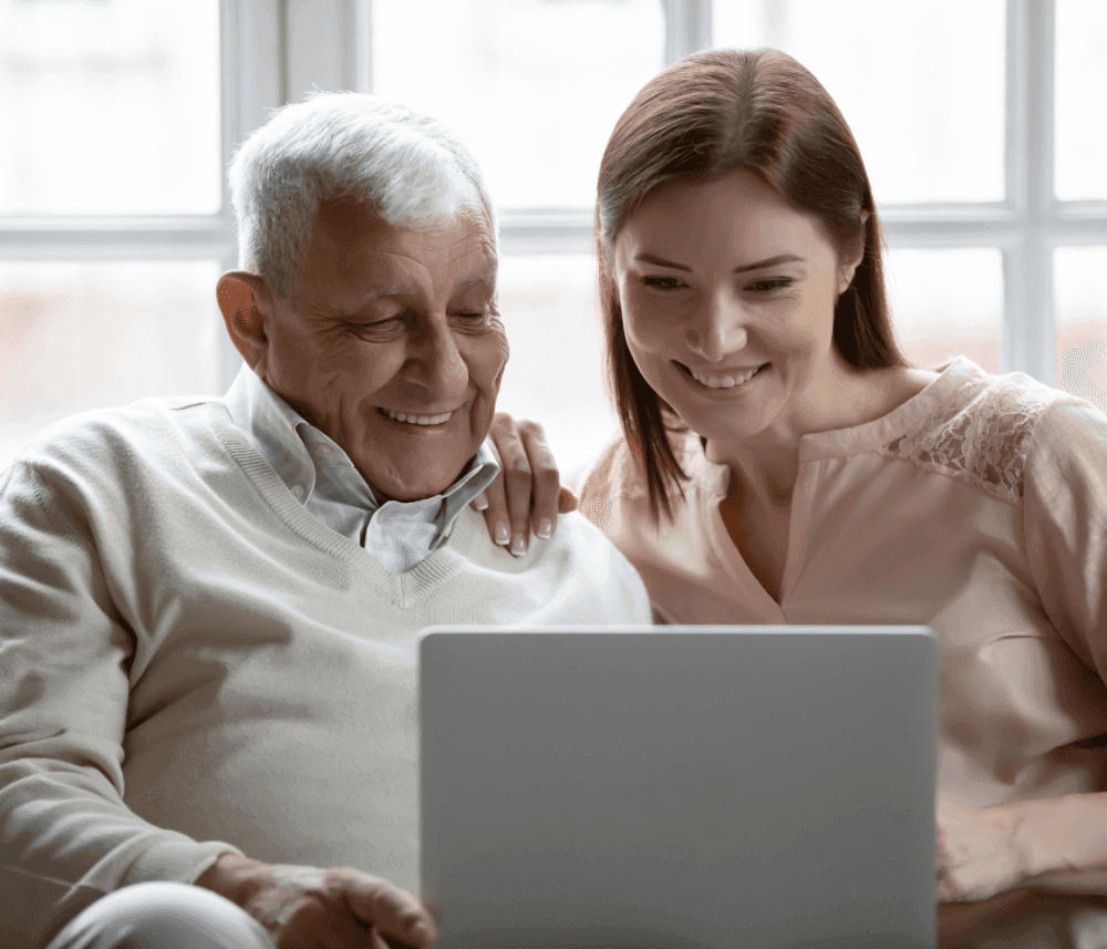 An older man and a younger woman smile while looking at a laptop screen, sitting together indoors by a window. - Home Instead