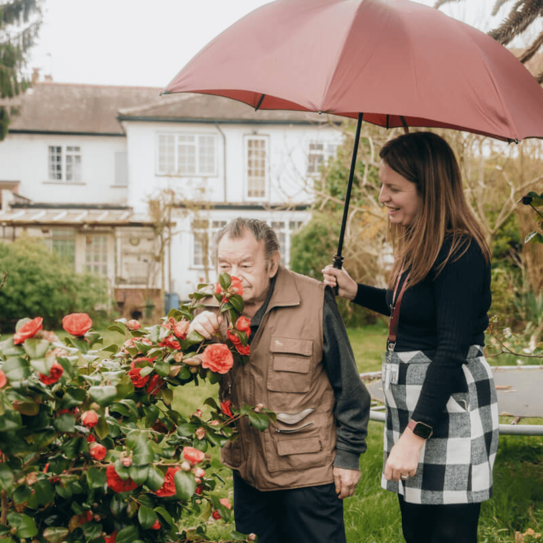 A woman holds a red umbrella over a man who is sniffing flowers in a garden near a house. - Home Instead