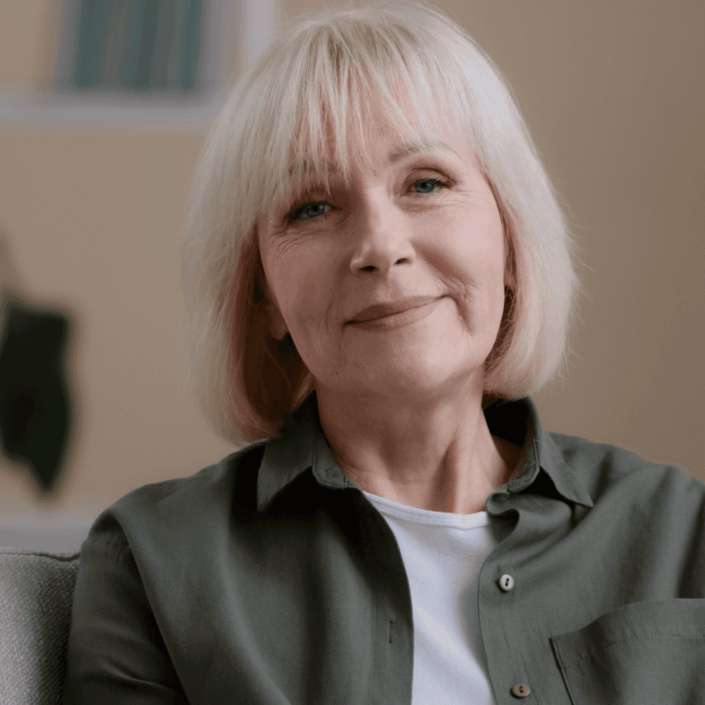 Older woman with short blond hair, smiling gently, wearing a green button-up shirt over a white top, indoors. - Home Instead
