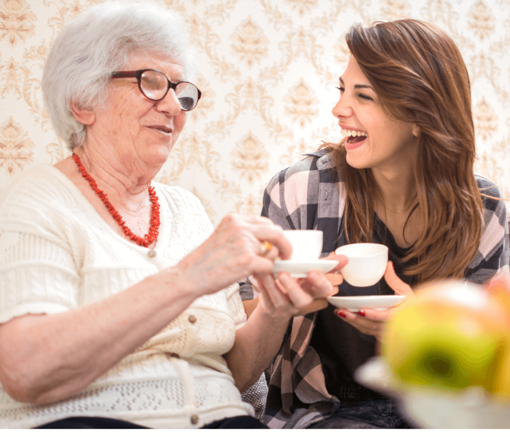 An elderly woman and a young woman share a laugh while holding cups of tea, sitting in a cozy, patterned room. - Home Instead