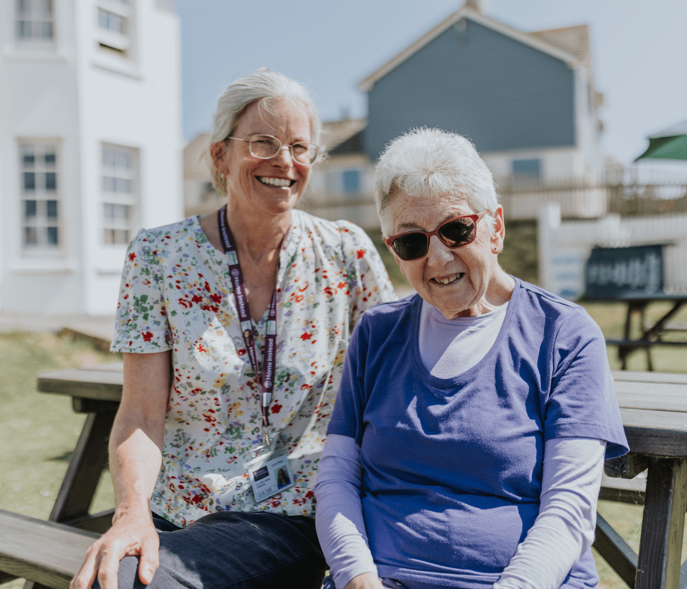 Two smiling elderly women sitting outdoors on a picnic bench with houses in the background. - Home Instead
