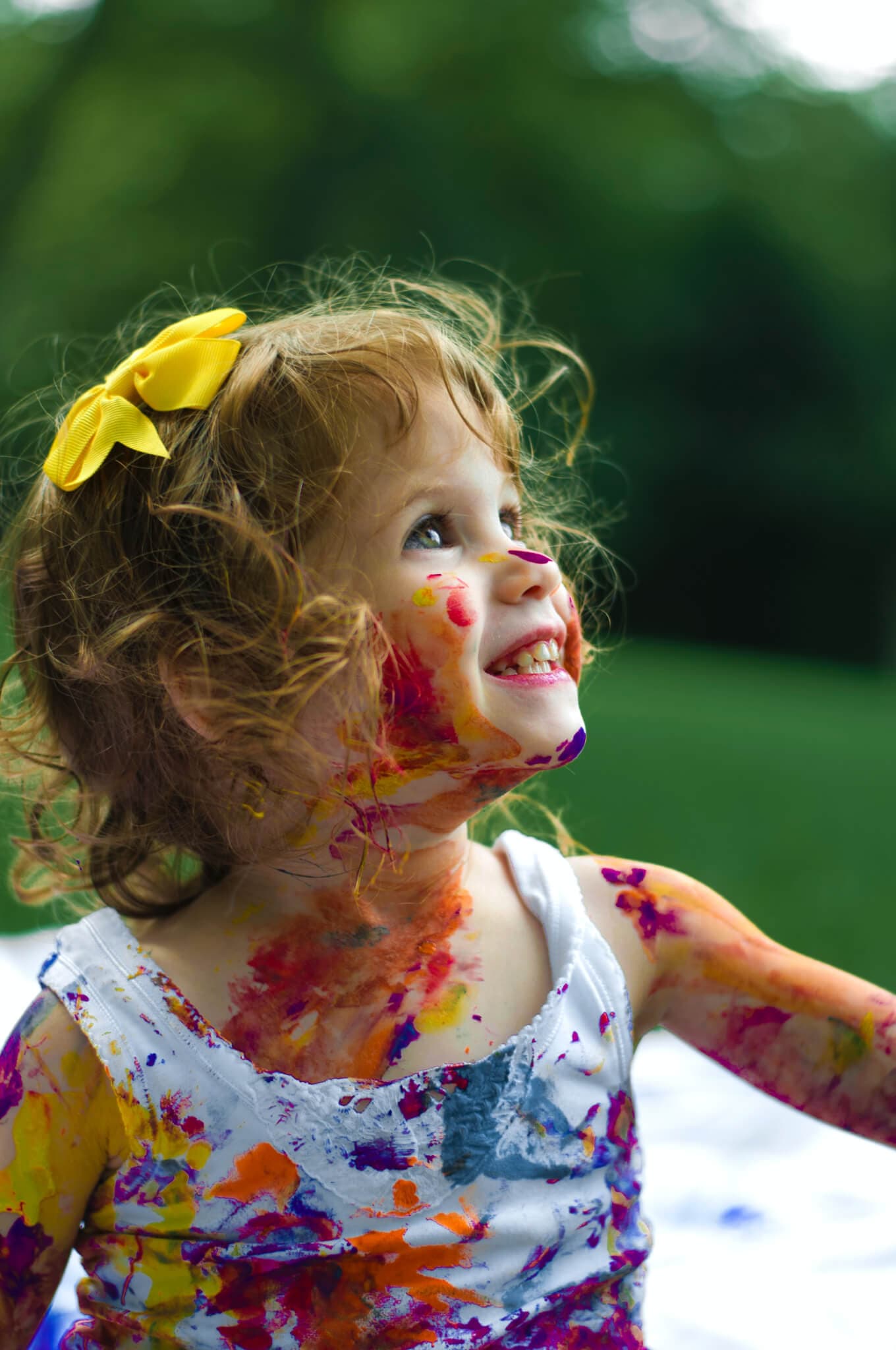 Smiling child with curly hair and a yellow bow, covered in colorful paint, sits on a blanket outdoors. - Home Instead