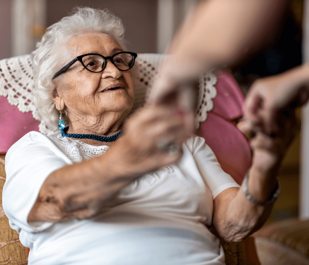 Elderly woman with glasses and a white blouse holding hands with an unseen person, smiling warmly in a cozy room. - Home Instead