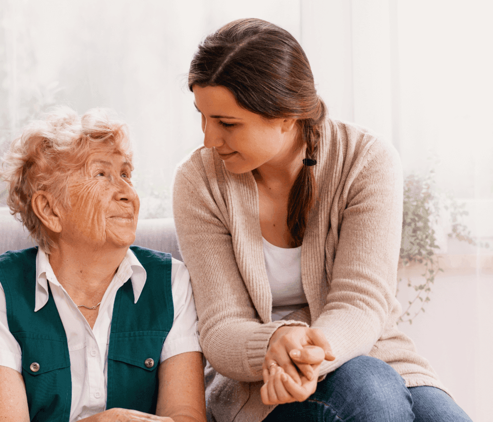 Young woman smiling at an elderly woman with love and care, sitting together in a bright room. - Home Instead