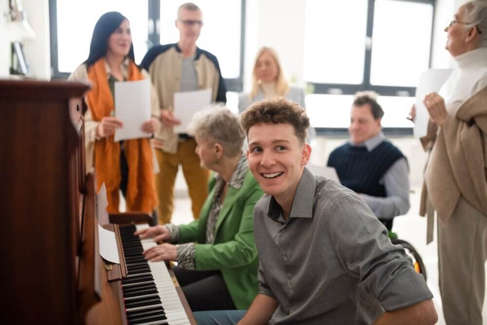 A young man sits at a piano, smiling, surrounded by people holding papers, possibly in a music or choir setting. - Home Instead