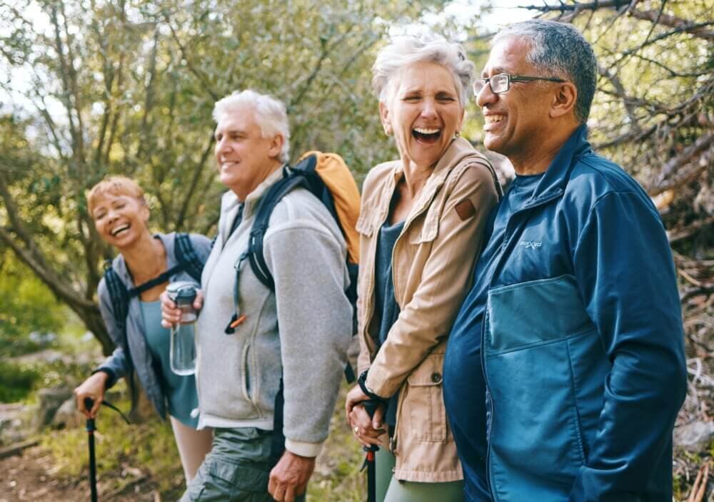 Four adults laughing and smiling while hiking in the woods, with trees and greenery in the background. - Home Instead