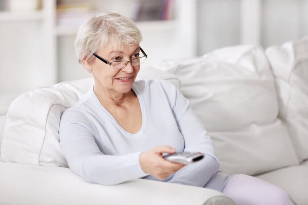 A smiling elderly woman with glasses holds a remote control while sitting on a white couch. - Home Instead