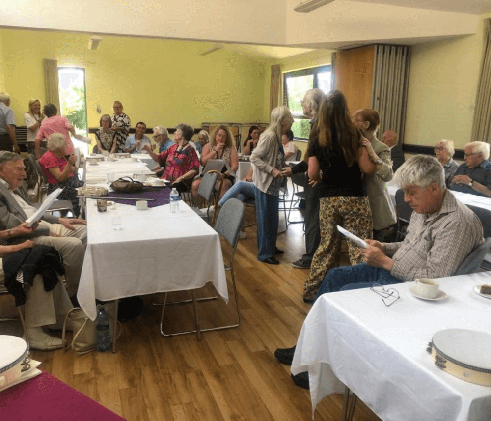 A group of people in a well-lit room, sitting and standing around tables with white cloths, engaged in conversation. - Home Instead