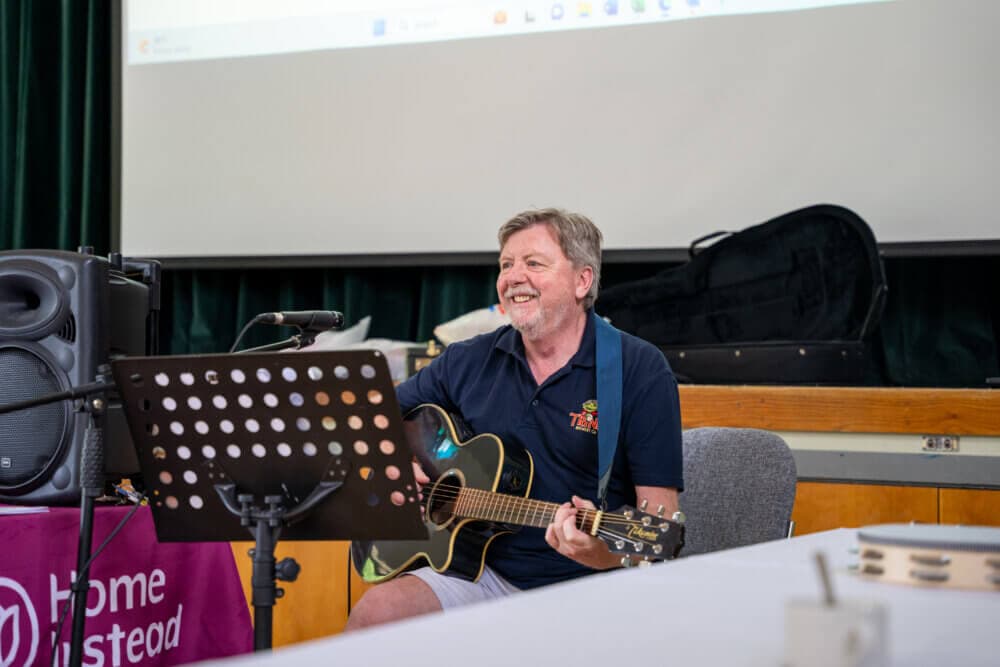 A man sits on a chair playing an acoustic guitar, smiling, with a microphone and a music stand in front of him. - Home Instead