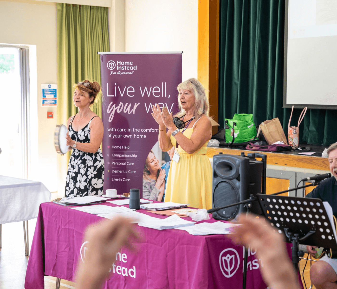 Two women stand clapping next to a banner in a decorated room, with a table displaying Home Instead materials in the foreground. - Home Instead