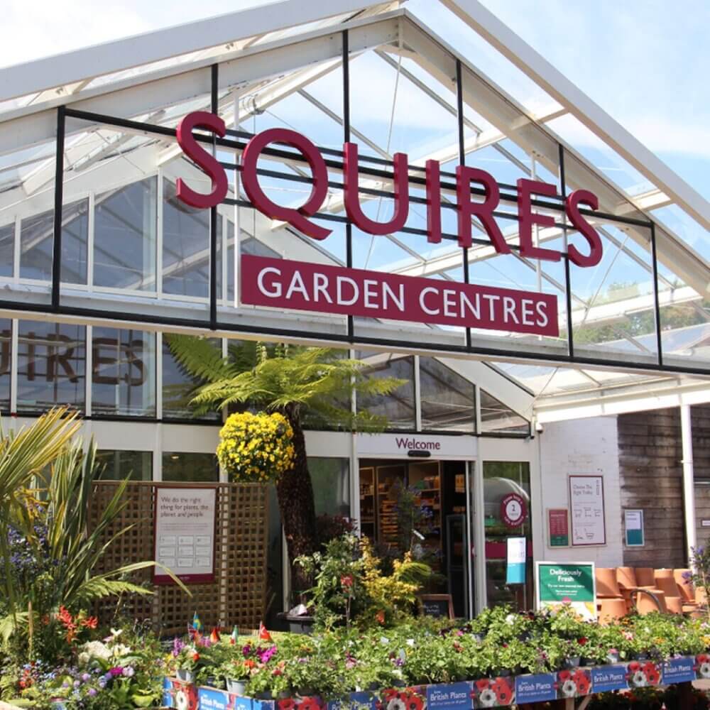 Entrance of Squires Garden Centres with plants and flowers displayed outside under a clear blue sky. - Home Instead