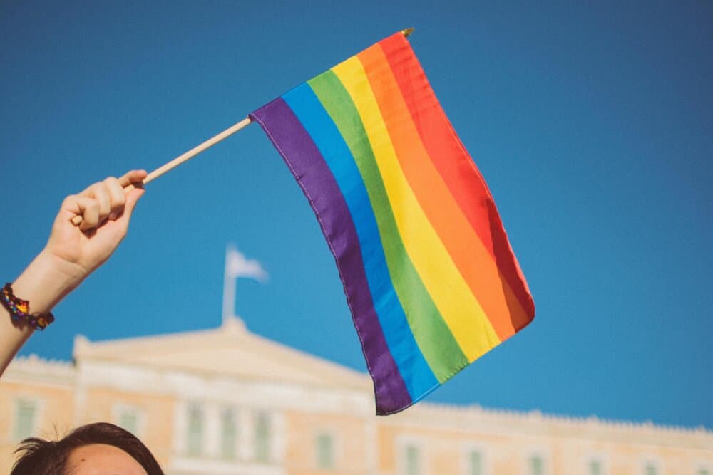 A person holding a rainbow flag against a clear blue sky with a building in the background. - Home Instead