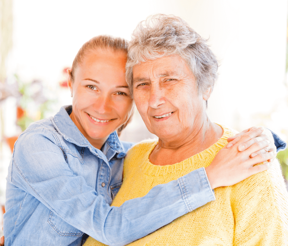 A young woman in a blue shirt smiles while hugging an older woman in a yellow sweater, both looking at the camera. - Home Instead