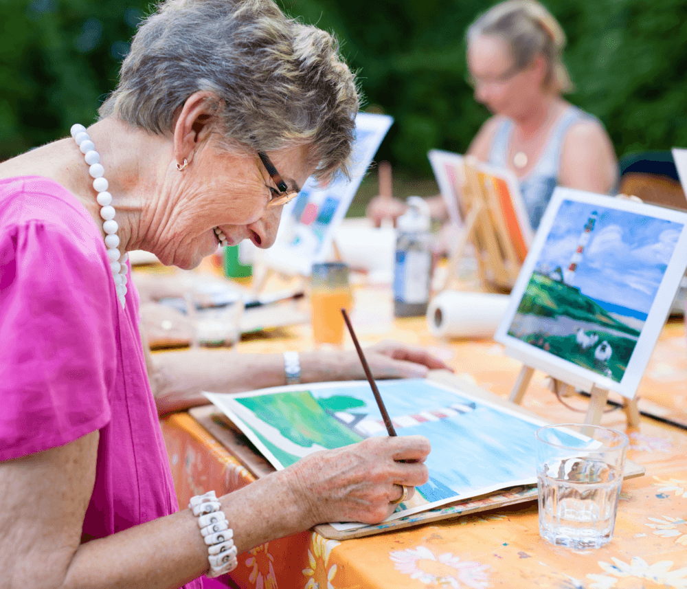 Elderly woman with white hair and glasses painting outdoors, surrounded by other people working on their own paintings. - Home Instead