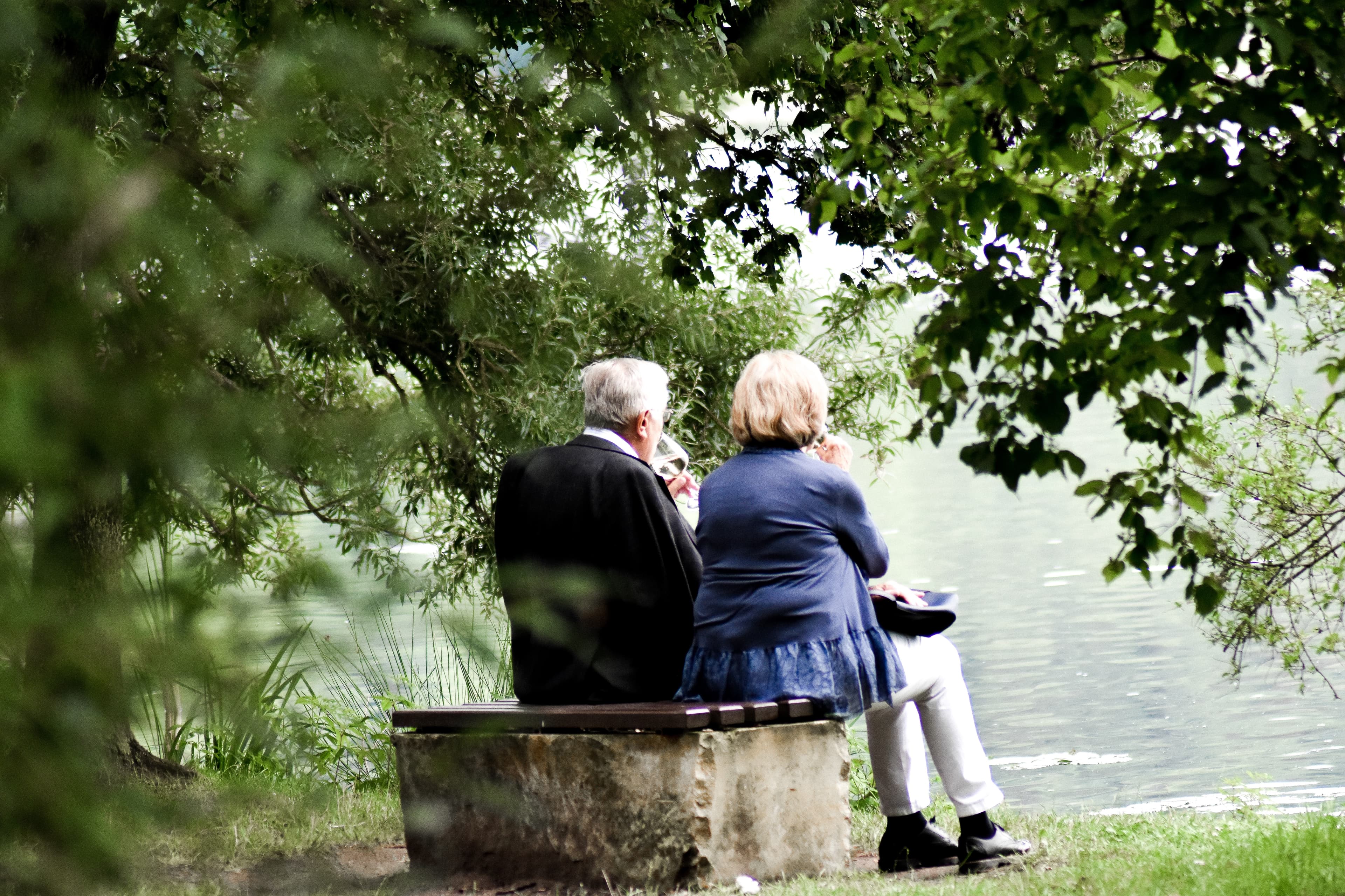 Two elderly people sit on a bench by a lake, surrounded by lush green trees, enjoying a serene moment outdoors. - Home Instead