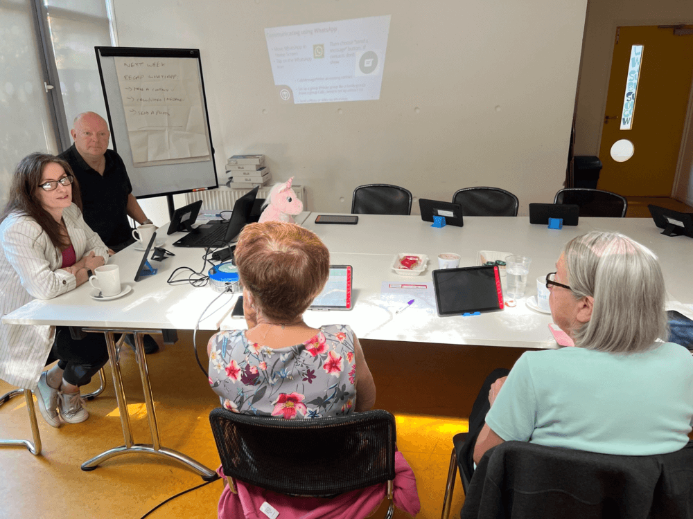 Four people sitting around a table with tablets and laptops, discussing something in a meeting room. - Home Instead