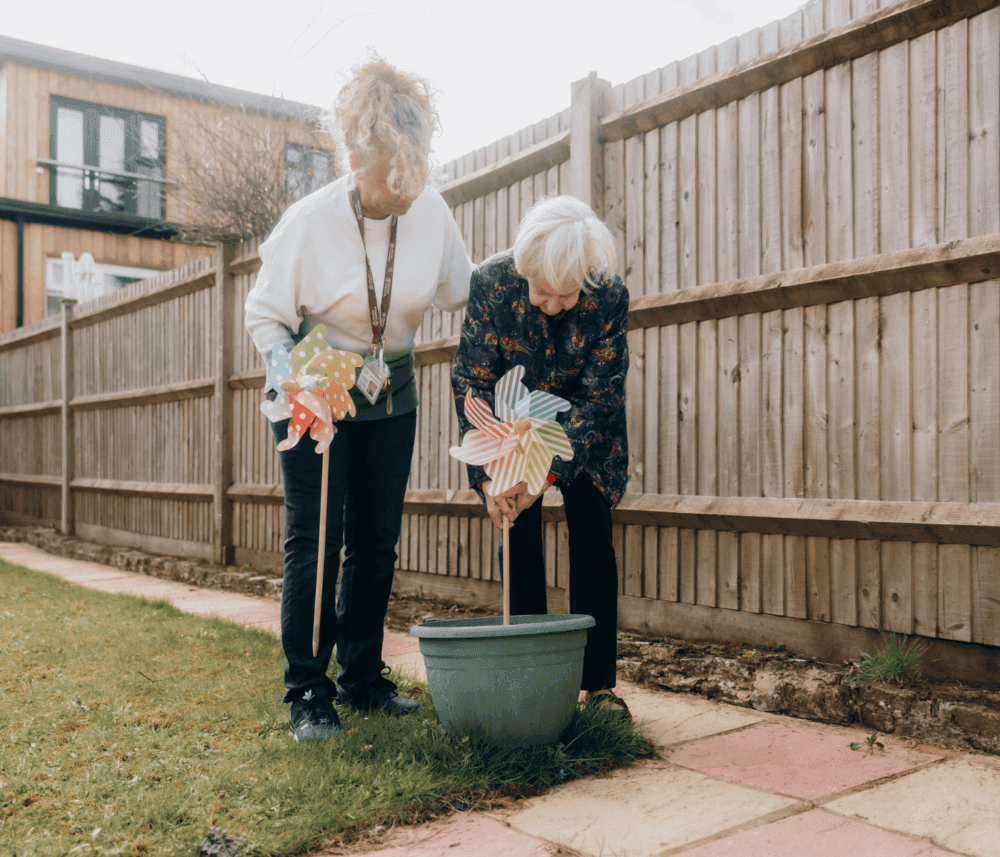 Two elderly women plant a pinwheel in a pot in a backyard garden, standing next to a wooden fence. - Home Instead