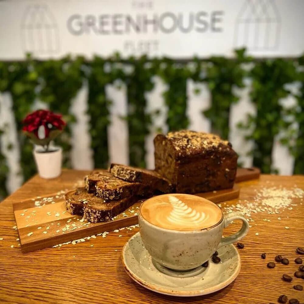 A latte and sliced cake on a wooden table at The Greenhouse Café, with green foliage and a red flower in the background. - Home Instead