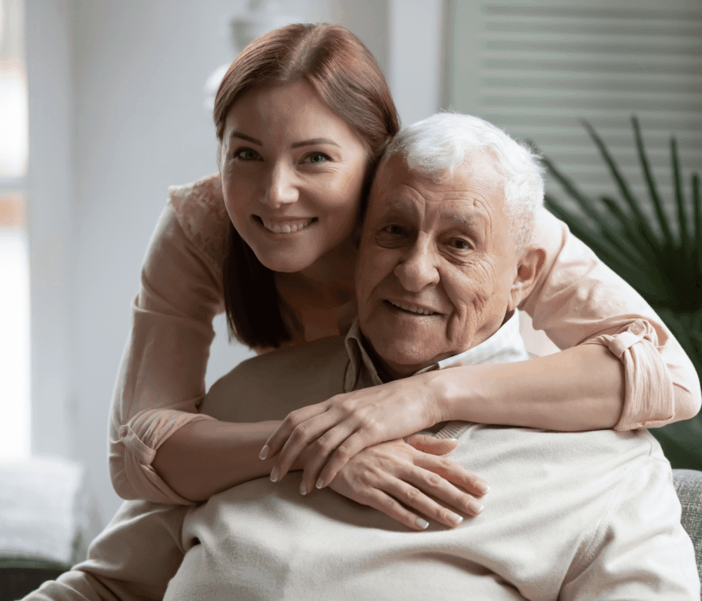 A young woman smiles and hugs a senior man from behind as they sit indoors, both looking at the camera. - Home Instead