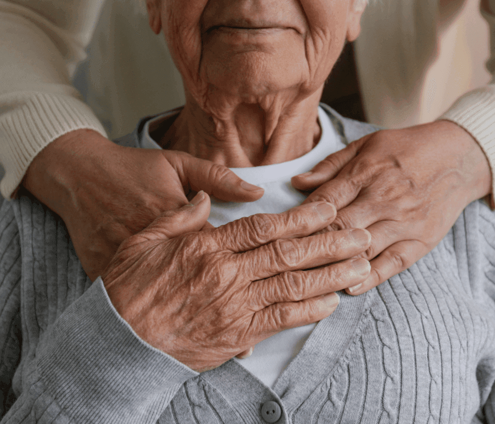 An elderly person with hands on their chest, embraced from behind by another person. - Home Instead