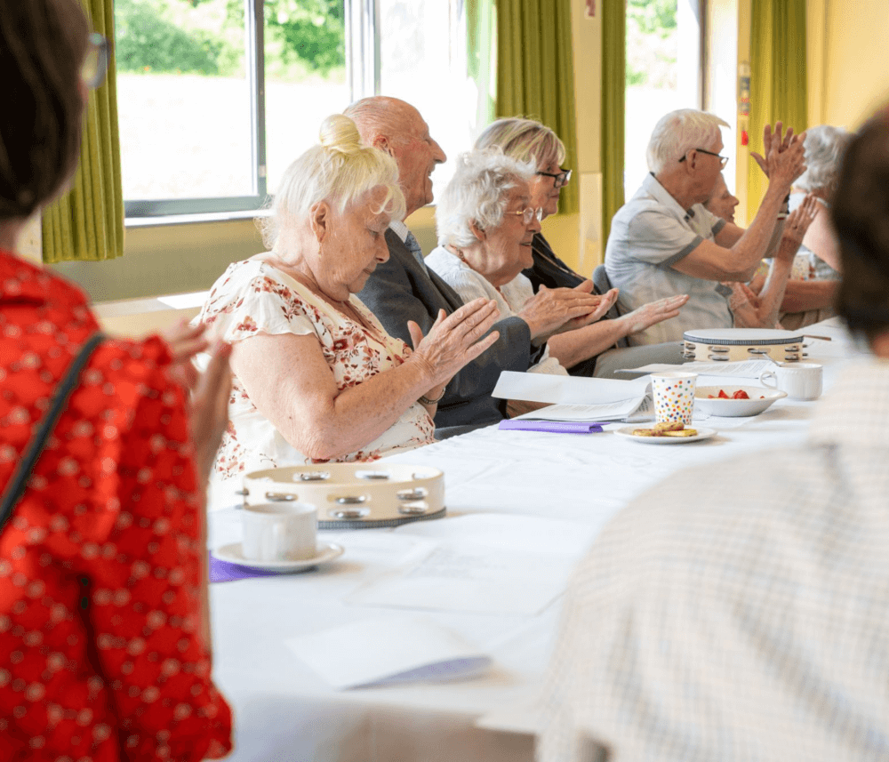 A group of elderly people clap their hands around a table with tambourines, tea cups, and documents in a bright room. - Home Instead