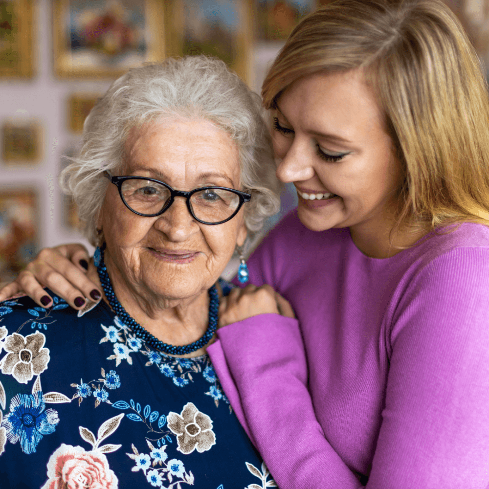 An elderly woman with glasses and a younger woman in a pink sweater smile and embrace indoors. - Home Instead