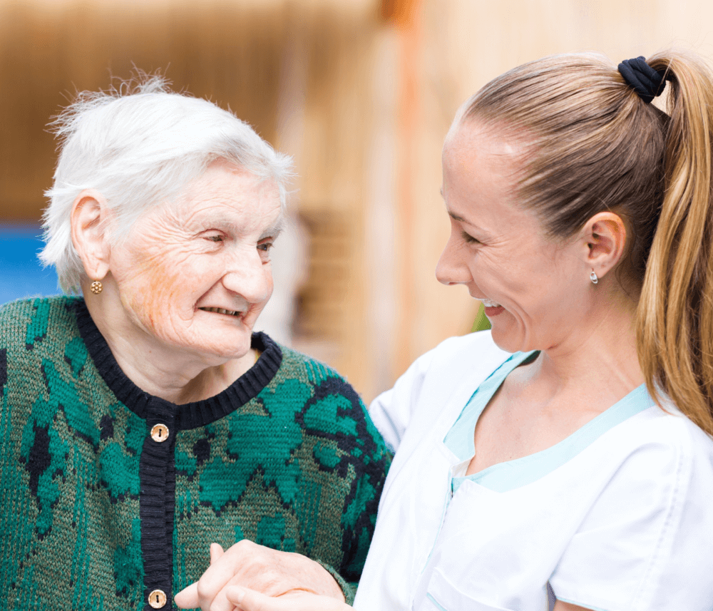 Elderly woman smiling at a caregiver who is wearing a white uniform and smiling back, holding onto each other’s hands. - Home Instead