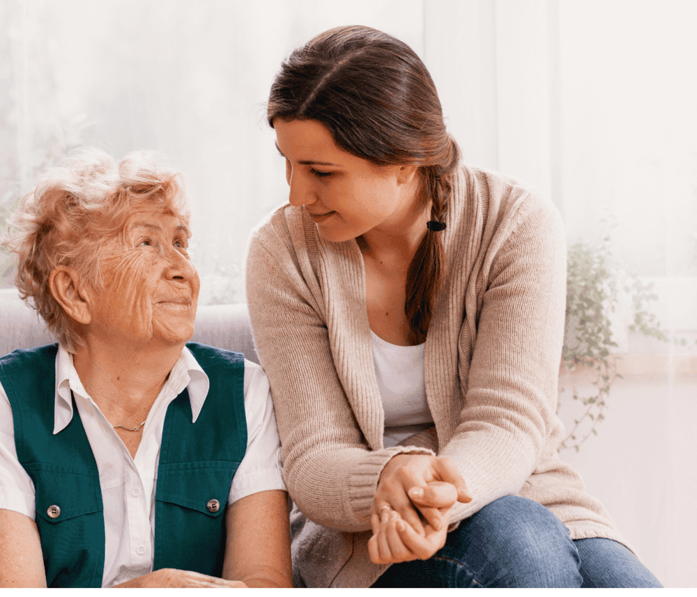An elderly woman and a younger woman sit together, smiling and looking at each other affectionately. - Home Instead