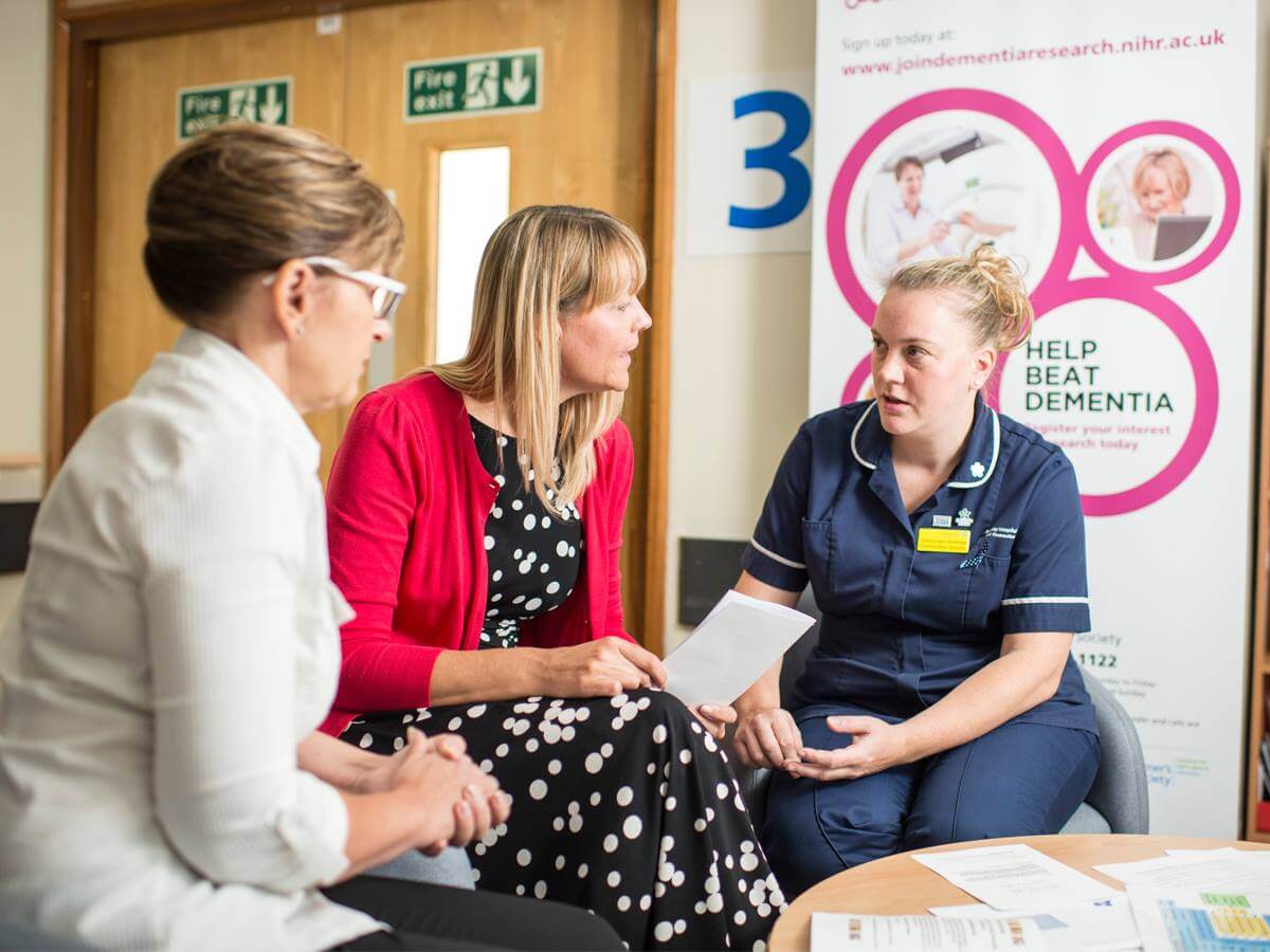 Three women having a discussion in a hospital setting, one wearing a nurse's uniform and the others in casual clothing. - Home Instead