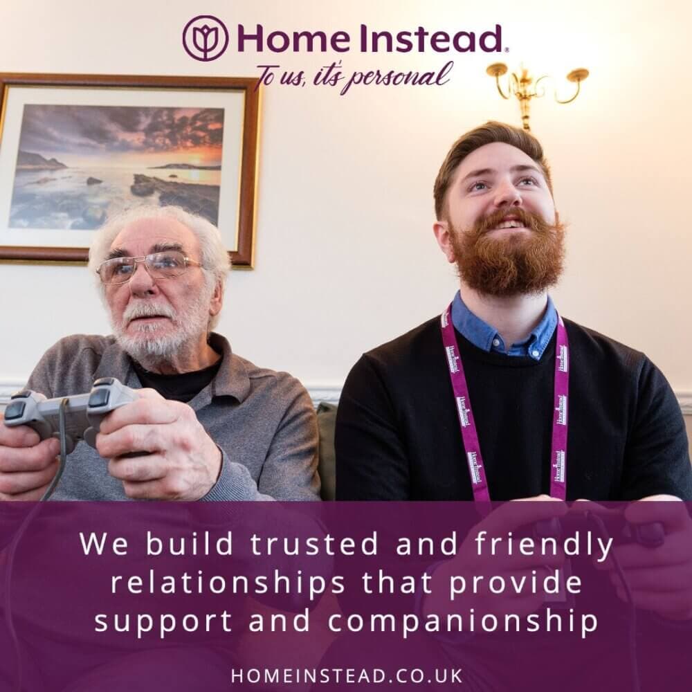 Two men, one older and one younger, playing video games with 'Home Instead' branding and text about support and companionship. - Home Instead