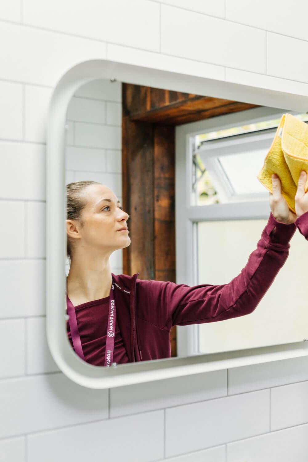 A person wearing a maroon shirt cleans a large bathroom mirror with a yellow cloth. - Home Instead