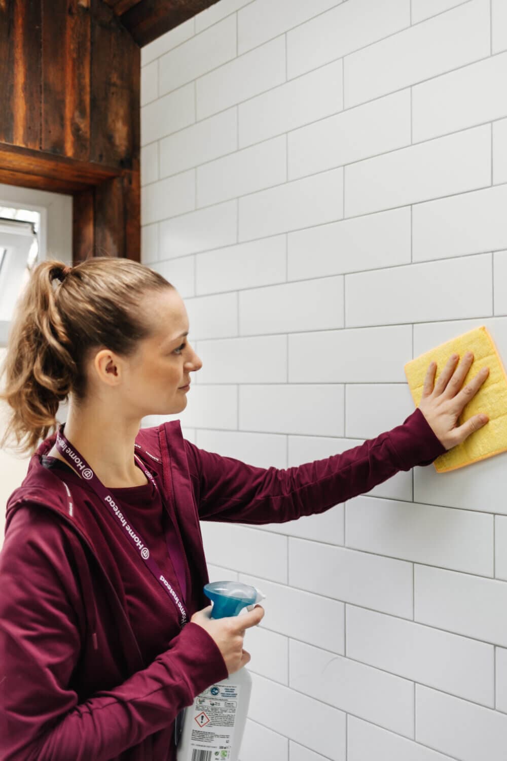 Person wearing a maroon top cleaning white tiled wall with a yellow sponge and holding a spray bottle. - Home Instead