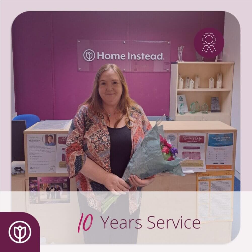 A woman stands in an office holding a bouquet, celebrating 10 years of service at Home Instead. - Home Instead