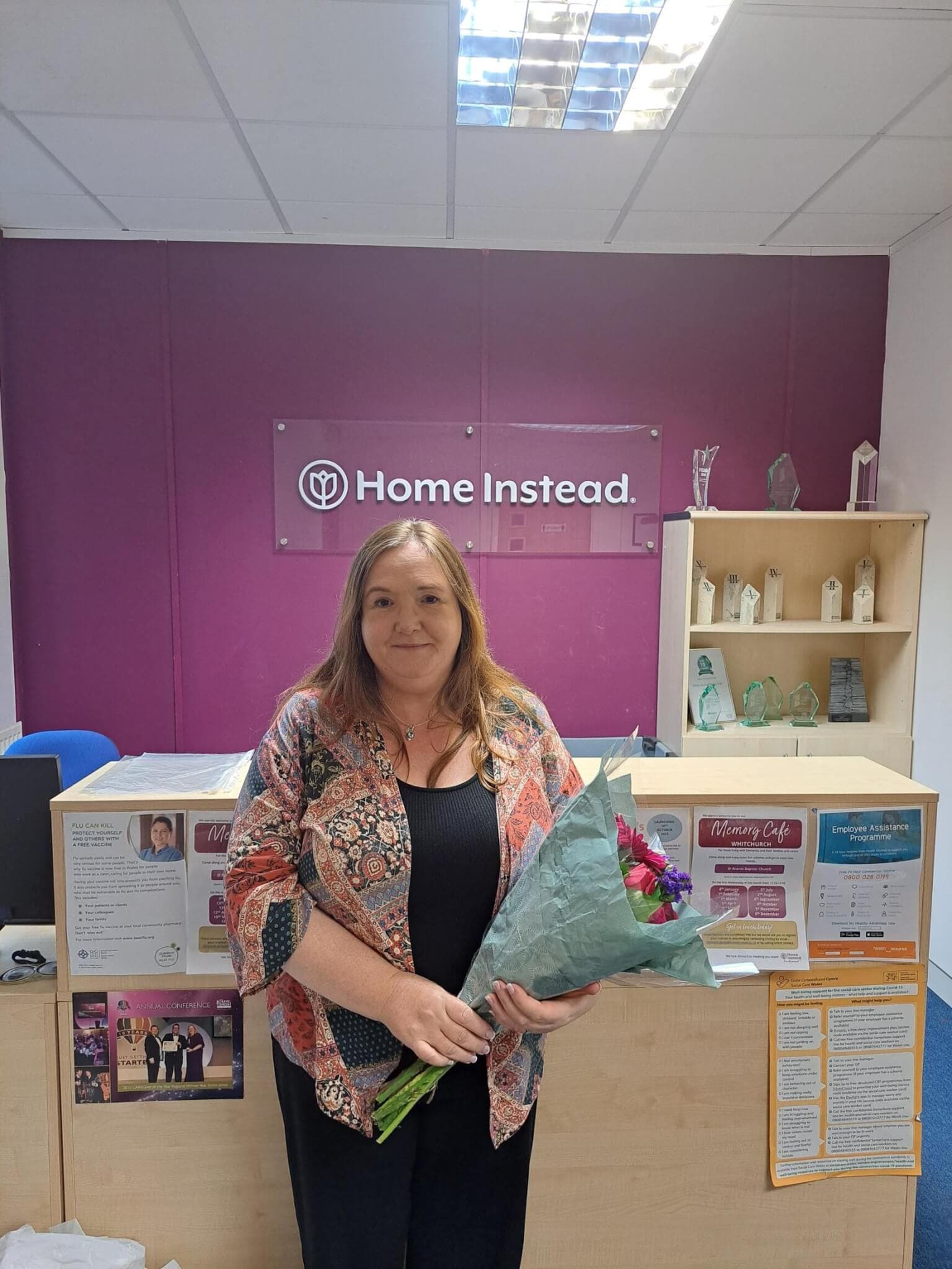 A woman stands holding a bouquet of flowers in an office with a "Home Instead." sign on the back wall. - Home Instead