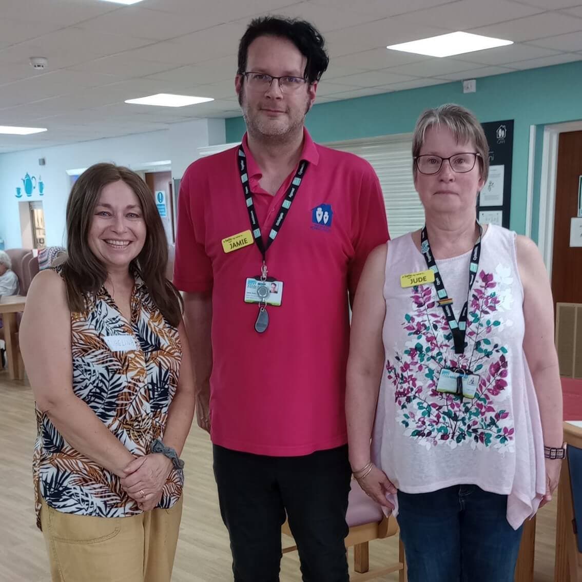 Three people stand together in a well-lit room, smiling and wearing name tags. - Home Instead