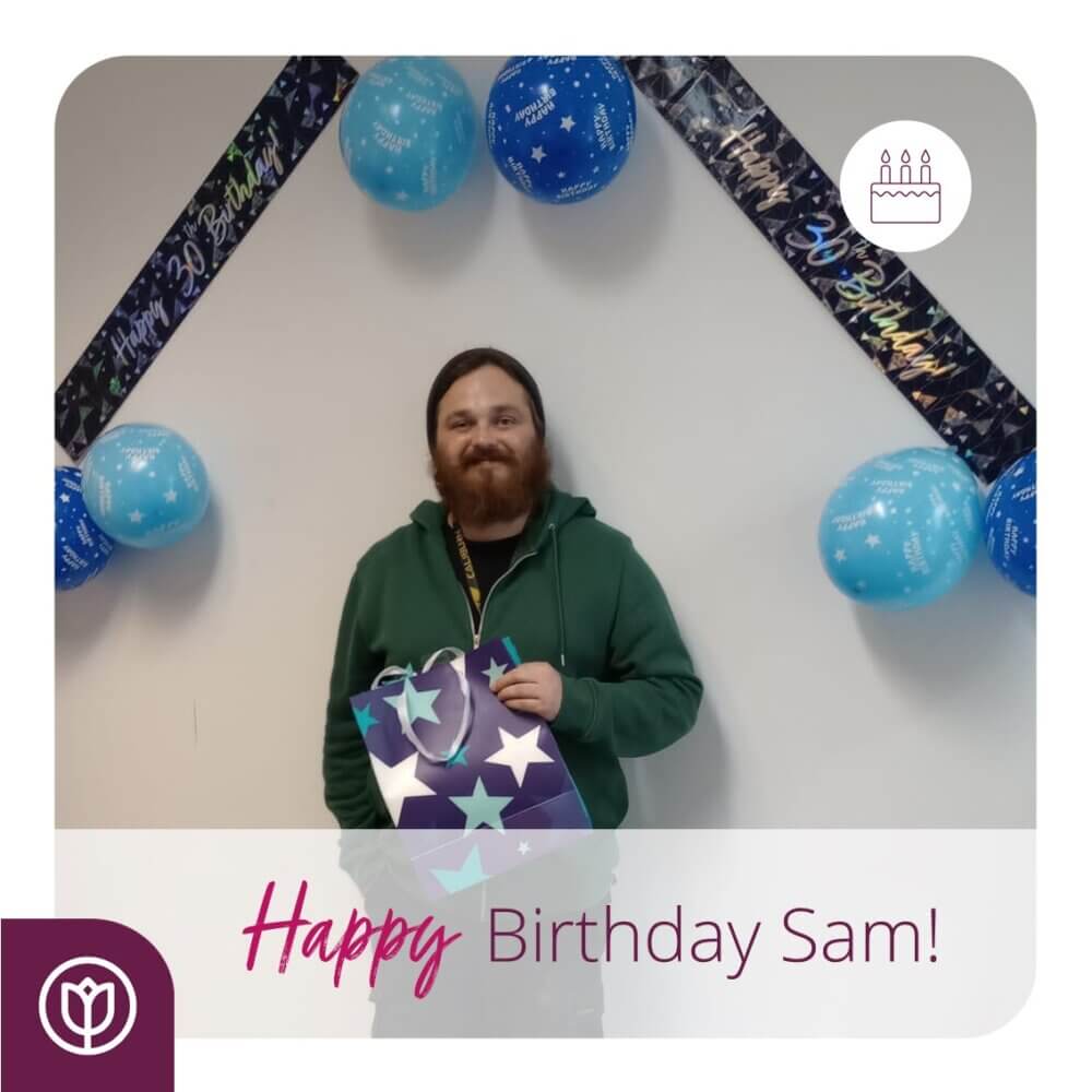 Man standing in front of birthday decorations, holding a gift bag. Text reads "Happy Birthday Sam!. - Home Instead