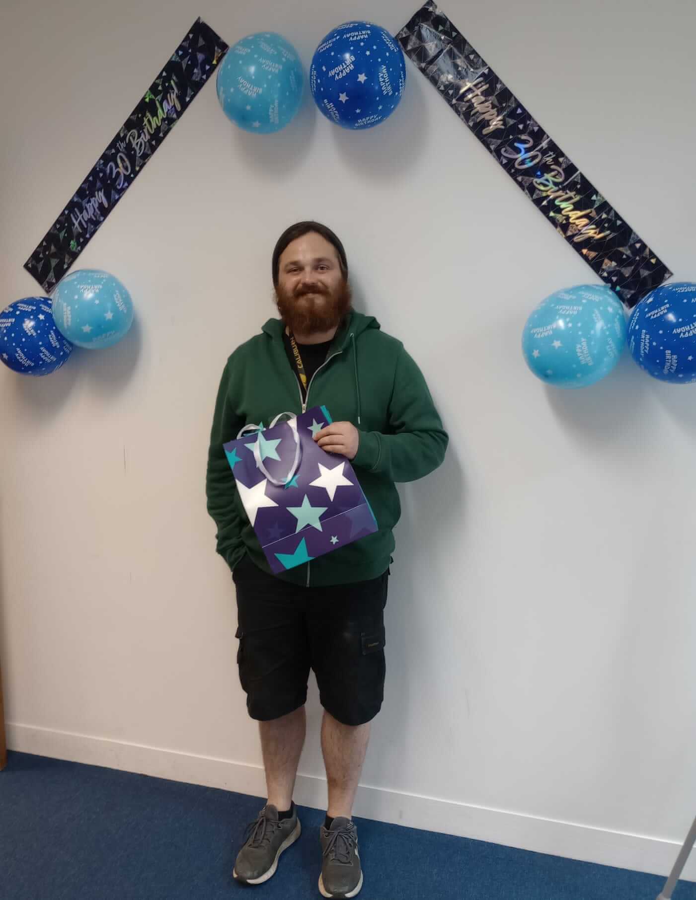 A person with a beard standing under birthday banners with blue balloons, holding a gift bag with star patterns. - Home Instead