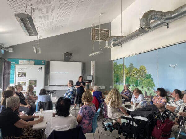 People seated at tables in a classroom, watching a presentation projected on a screen. - Home Instead