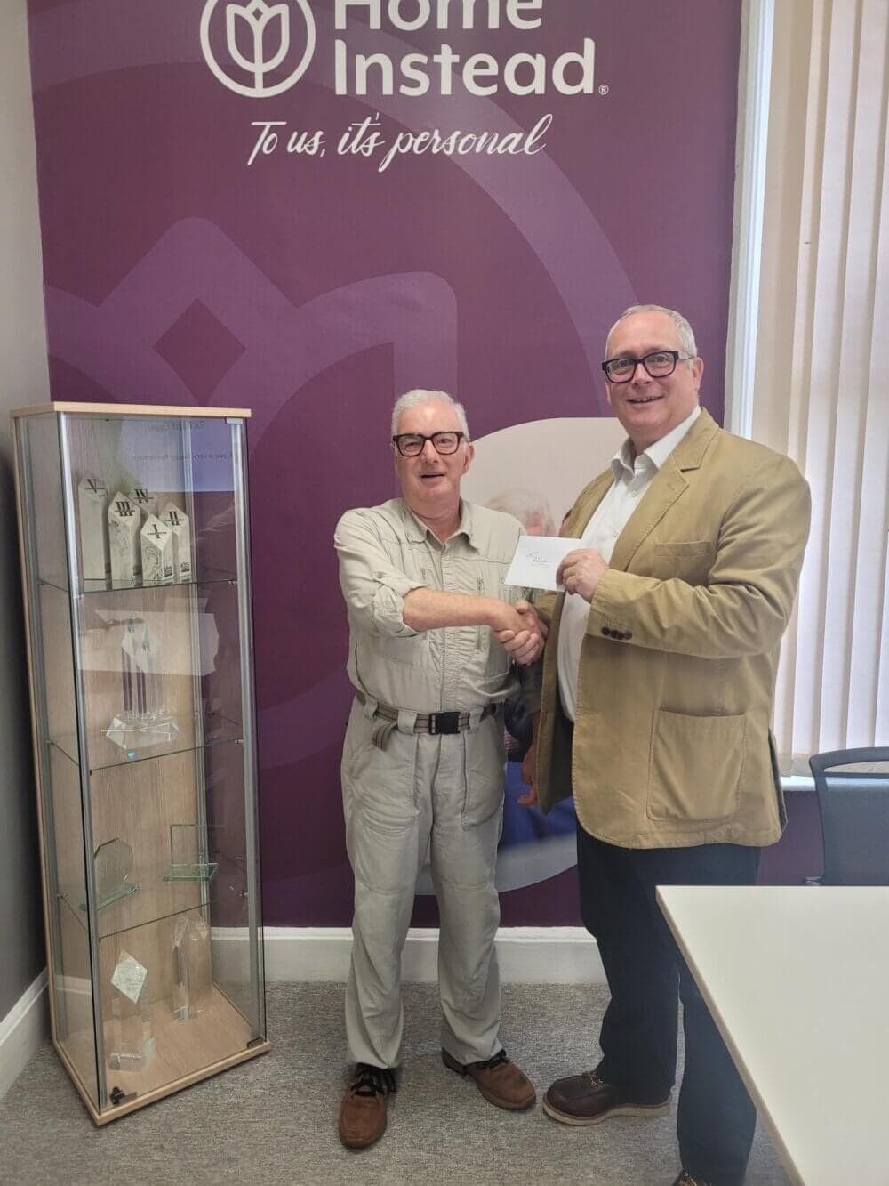 Two men shaking hands in an office with a "Home Instead" sign in the background. - Home Instead