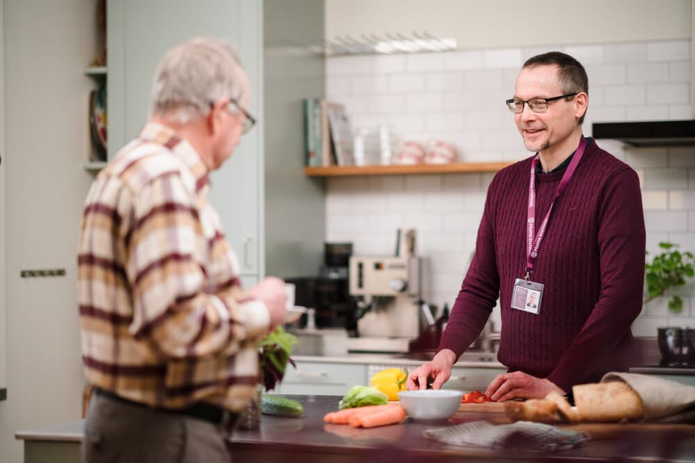 Two men having a conversation in a kitchen, one chopping vegetables and the other holding a mug. - Home Instead