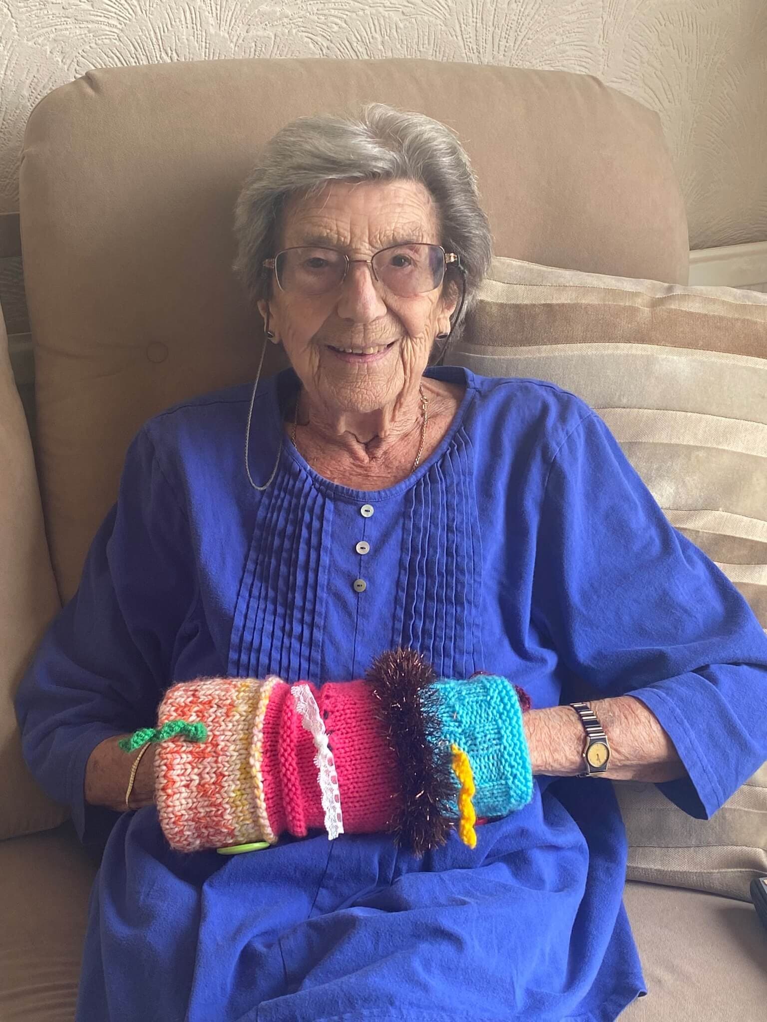 Elderly woman with glasses in a blue dress, sitting on a couch, holding colorful knitted items. - Home Instead