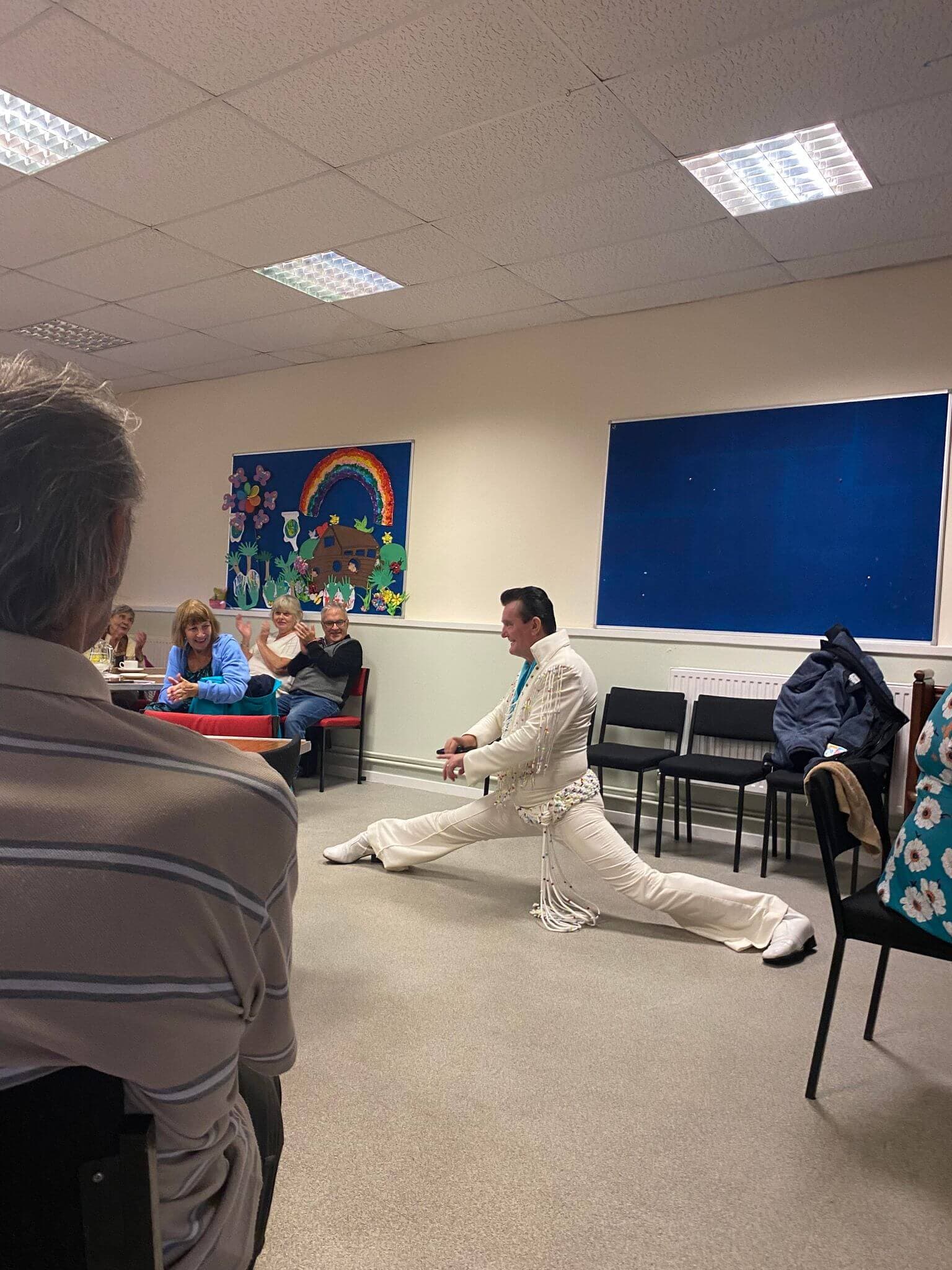 A performer in a white jumpsuit does the splits in a room with seated spectators and colorful art on the wall. - Home Instead