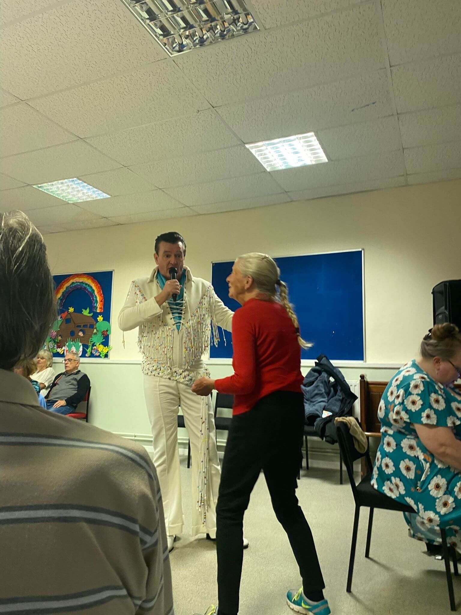 Performer in white outfit singing into a microphone while a woman in red dances in a well-lit room. - Home Instead